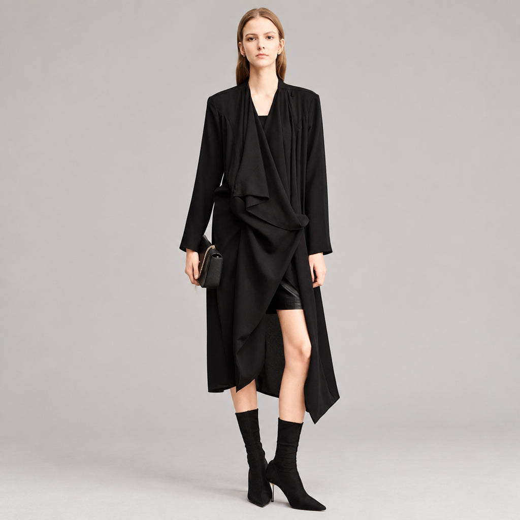 A model wearing a long black draped wrap coat with long sleeves, styled with black boots and holding a small black clutch, set against a minimalist studio background with the AMII logo in the top left corner.