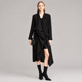 A model wearing a long black draped wrap coat with long sleeves, styled with black boots and holding a small black clutch, set against a minimalist studio background with the AMII logo in the top left corner.