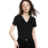 A woman modeling a black AMII short-sleeve V-neck top with a twisted knot detail at the chest, paired with black pants and a black belt with a gold buckle, against a light beige background.