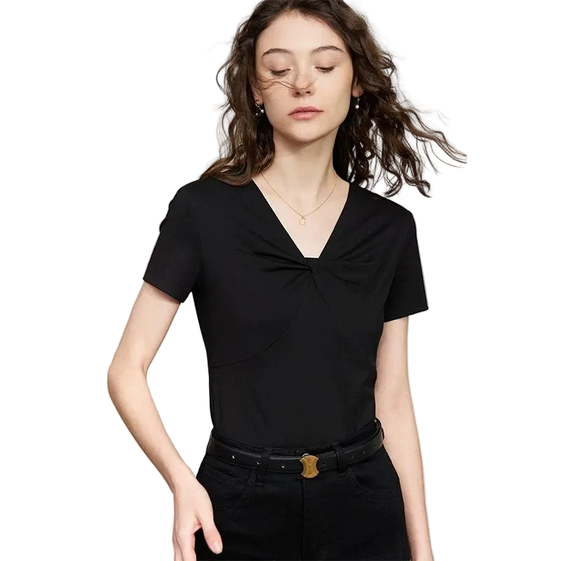 A woman modeling a black AMII short-sleeve V-neck top with a twisted knot detail at the chest, paired with black pants and a black belt with a gold buckle, against a light beige background.